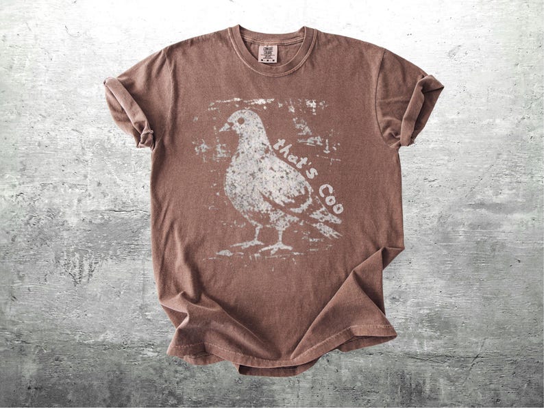 May include: Brown t-shirt with a distressed white graphic of a pigeon and the text "that's Coo". The shirt has rolled-up sleeves and a crew neck. The shirt is made of a soft material.