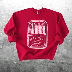 May include: A red sweatshirt with a white graphic of a sardine can. The can contains four fish and the words "Sardines Wild Caught in Olive Oil". The sweatshirt has cuffed sleeves.