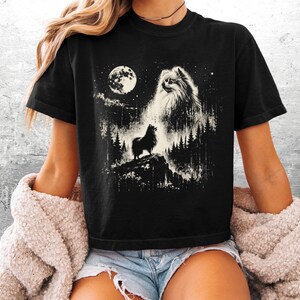 May include: A black short-sleeve t-shirt featuring a white graphic print of a fluffy Pomeranian dog standing on a rocky cliff in a forest at night. The design includes a large full moon, starry sky, and a ghostly, ethereal image of the dog's face.