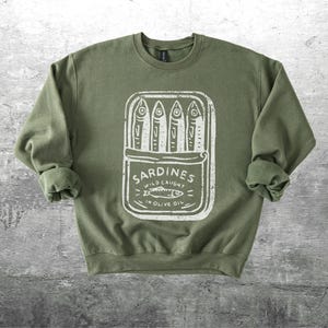 May include: Olive green sweatshirt with a white graphic of a sardine can. The can design includes the word "Sardines" and the text "Wild Caught in Olive Oil". The sweatshirt has a crew neck and long sleeves.