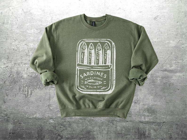 May include: Olive green sweatshirt with a white graphic of a sardine can. The can design includes the word "Sardines" and the text "Wild Caught in Olive Oil". The sweatshirt has a crew neck and long sleeves.