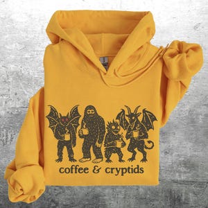 May include: Mustard yellow hoodie with a graphic of cryptids holding coffee mugs. The cryptids include a bat-like creature, Bigfoot, a horned demon, and a small, spiky creature. The text "coffee & cryptids" is below the graphic.