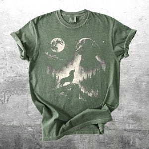 May include: Olive green t-shirt featuring a graphic of a dachshund dog silhouette, a full moon, and a forest scene. The design is printed in shades of gray and white. The shirt has short sleeves and a crew neck.