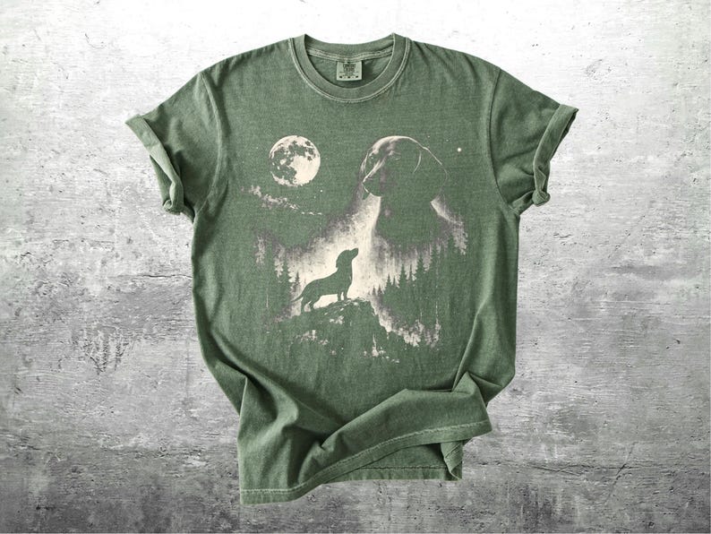 May include: Olive green t-shirt featuring a graphic of a dachshund dog silhouette, a full moon, and a forest scene. The design is printed in shades of gray and white. The shirt has short sleeves and a crew neck.