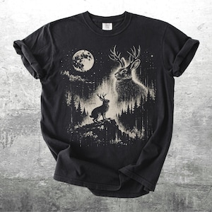 May include: Black t-shirt featuring a detailed illustration of a jackalope with antlers, set against a forest and a full moon. The design is in shades of white and beige, creating a striking contrast on the dark fabric.