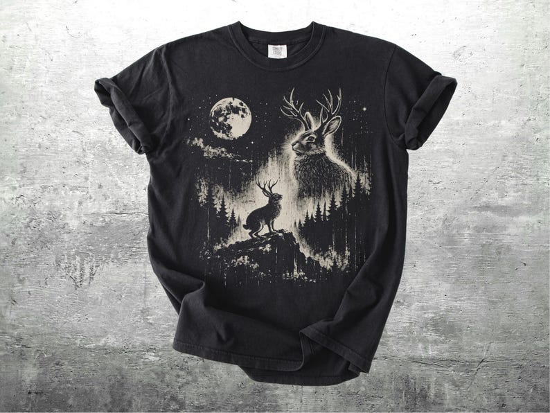 May include: Black t-shirt featuring a detailed illustration of a jackalope with antlers, set against a forest and a full moon. The design is in shades of white and beige, creating a striking contrast on the dark fabric.
