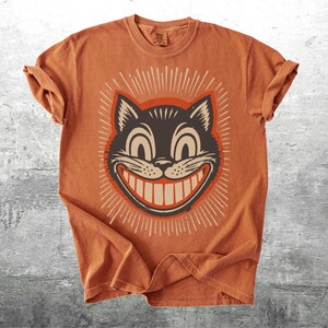 Op de afbeelding: Een kortmouwig T-shirt in gebrand oranje met een grafische print van een zwart kattengezicht met een brede, tandige grijns en grote ogen. De kattenillustratie is omringd door een stralend patroon van dunne witte lijnen op de borst.