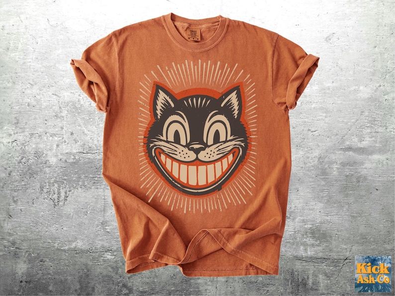 Op de afbeelding: Een kortmouwig T-shirt in gebrand oranje met een grafische print van een zwart kattengezicht met een brede, tandige grijns en grote ogen. De kattenillustratie is omringd door een stralend patroon van dunne witte lijnen op de borst.