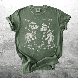 K&ouml;nnte beinhalten: Gr&uuml;nes T-Shirt mit einem verblassten Aufdruck von zwei Punkrock-Ratten, jede mit Irokesenschnitt und Nietenhalsband. Die Ratten blicken sich an. Das T-Shirt hat gerollte &Auml;rmel und einen Rundhalsausschnitt. Der Stoff wirkt weich und bequem.