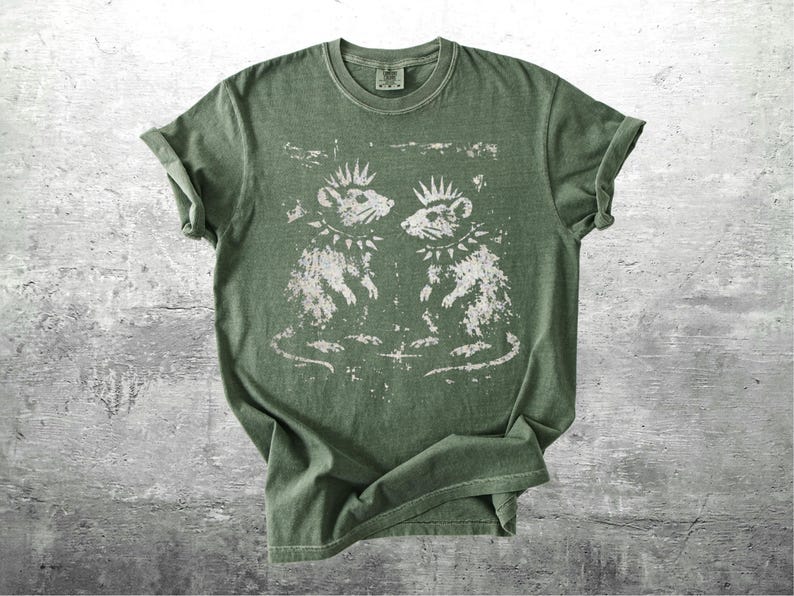 K&ouml;nnte beinhalten: Gr&uuml;nes T-Shirt mit einem verblassten Aufdruck von zwei Punkrock-Ratten, jede mit Irokesenschnitt und Nietenhalsband. Die Ratten blicken sich an. Das T-Shirt hat gerollte &Auml;rmel und einen Rundhalsausschnitt. Der Stoff wirkt weich und bequem.