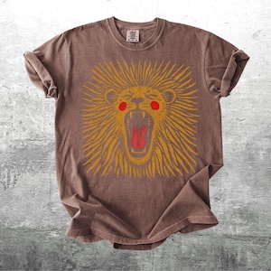 May include: Brown t-shirt with a roaring lion design. The lion has a yellow mane, red mouth, and red cheeks. The shirt is made of a soft material and has rolled-up sleeves.