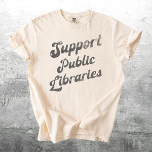 May include: Cream-colored t-shirt with the text "Support Public Libraries" in a distressed, dark gray font. The shirt has rolled-up sleeves and a classic crew neck design. The shirt is made of a soft material.