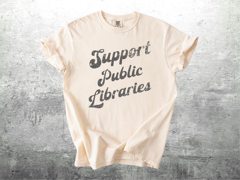 May include: Cream-colored t-shirt with the text "Support Public Libraries" in a distressed, dark gray font. The shirt has rolled-up sleeves and a classic crew neck design. The shirt is made of a soft material.