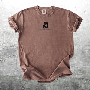 May include: Brown t-shirt with a small black and white cat graphic and the text "are you kitten me". The shirt has rolled-up sleeves and a crew neck. The shirt is made of a soft material.