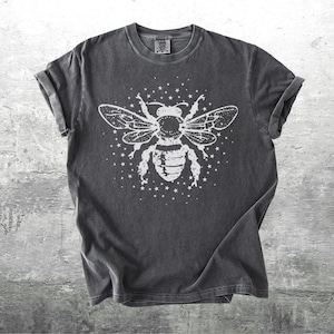 May include: Dark gray t-shirt featuring a large white graphic of a bee with outstretched wings and surrounding stars. The tee has rolled-up sleeves and a crew neck. The shirt is made of a soft, comfortable material.