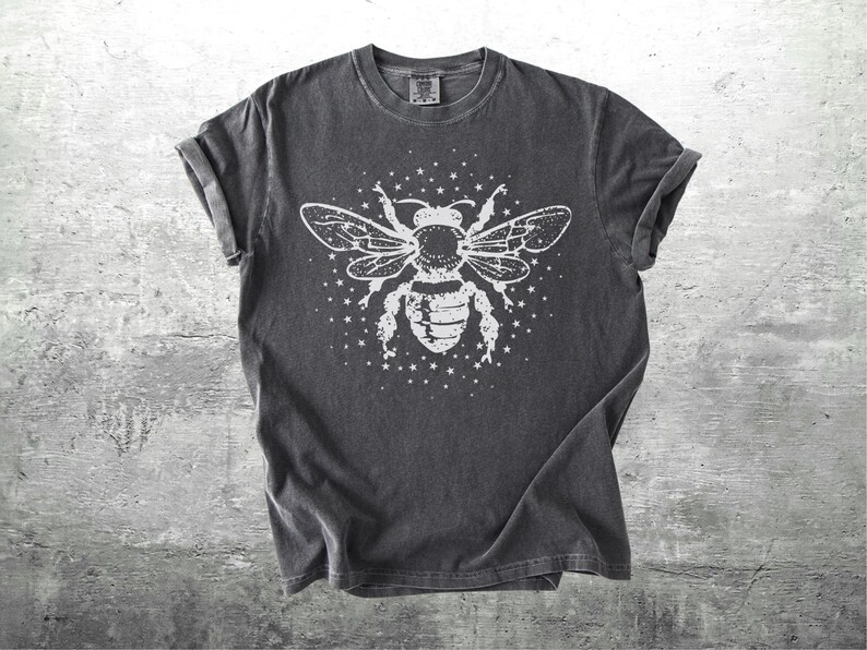 May include: Dark gray t-shirt featuring a large white graphic of a bee with outstretched wings and surrounding stars. The tee has rolled-up sleeves and a crew neck. The shirt is made of a soft, comfortable material.