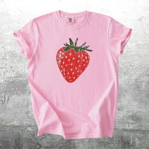 May include: A light pink t-shirt featuring a large, distressed red strawberry graphic with white seeds and green leaves. The shirt has short sleeves and a crew neck, perfect for casual wear.