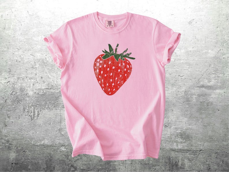 May include: A light pink t-shirt featuring a large, distressed red strawberry graphic with white seeds and green leaves. The shirt has short sleeves and a crew neck, perfect for casual wear.