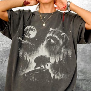 K&ouml;nnte beinhalten: Ein anthrazitfarbenes Kurzarm-T-Shirt mit einem Grafikdruck eines Waschb&auml;ren, eines Vollmonds und einer Waldlandschaft. Das Design zeigt ein gro&szlig;es Waschb&auml;rgesicht und einen kleineren Waschb&auml;ren, der auf einer felsigen Klippe l&auml;uft.