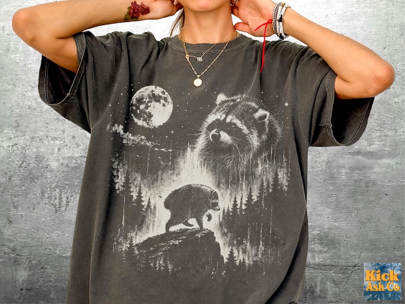 K&ouml;nnte beinhalten: Ein anthrazitfarbenes Kurzarm-T-Shirt mit einem Grafikdruck eines Waschb&auml;ren, eines Vollmonds und einer Waldlandschaft. Das Design zeigt ein gro&szlig;es Waschb&auml;rgesicht und einen kleineren Waschb&auml;ren, der auf einer felsigen Klippe l&auml;uft.