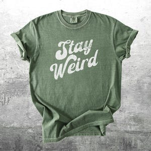 K&ouml;nnte beinhalten: Olivgr&uuml;nes T-Shirt mit dem wei&szlig;en Schriftzug "Stay Weird" in kursiver Schrift. Das T-Shirt hat hochgekrempelte &Auml;rmel und ein Vintage-Aussehen mit leicht verblichenen Farben. Das T-Shirt wird auf einer strukturierten, grauen Oberfl&auml;che pr&auml;sentiert.