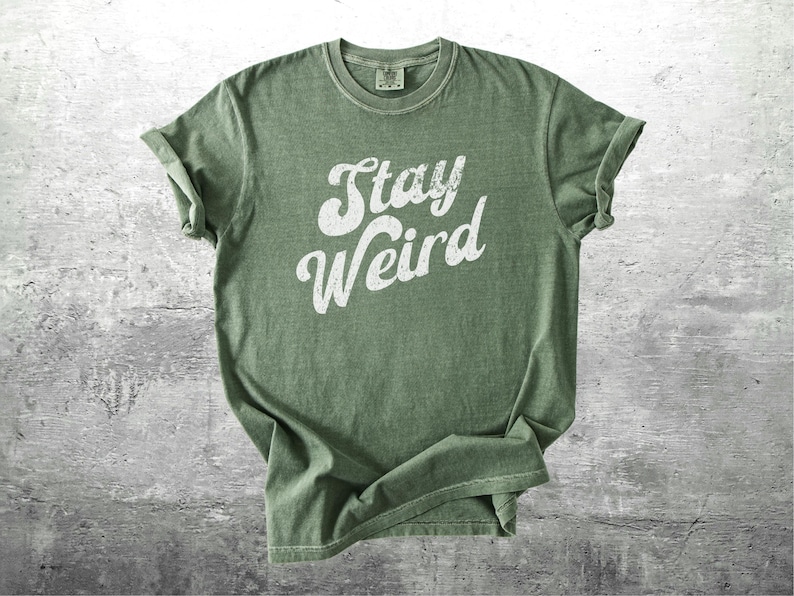 K&ouml;nnte beinhalten: Olivgr&uuml;nes T-Shirt mit dem wei&szlig;en Schriftzug "Stay Weird" in kursiver Schrift. Das T-Shirt hat hochgekrempelte &Auml;rmel und ein Vintage-Aussehen mit leicht verblichenen Farben. Das T-Shirt wird auf einer strukturierten, grauen Oberfl&auml;che pr&auml;sentiert.
