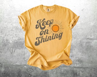 May include: Mustard yellow t-shirt with rolled sleeves. The shirt features the text "Keep on Shining" in a distressed font, with a sun graphic. The shirt is laid flat on a gray surface.