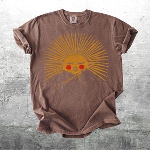 K&ouml;nnte beinhalten: Braunes T-Shirt mit einem Sonnen-Motiv, das ein l&auml;chelndes Gesicht und eine Bergkette zeigt. Die Sonne hat gelbe Strahlen und eine rote Wange. Das Shirt ist aus weichem Material und hat hochgekrempelte &Auml;rmel.