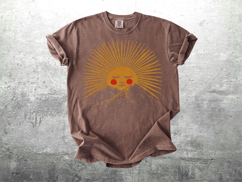 K&ouml;nnte beinhalten: Braunes T-Shirt mit einem Sonnen-Motiv, das ein l&auml;chelndes Gesicht und eine Bergkette zeigt. Die Sonne hat gelbe Strahlen und eine rote Wange. Das Shirt ist aus weichem Material und hat hochgekrempelte &Auml;rmel.