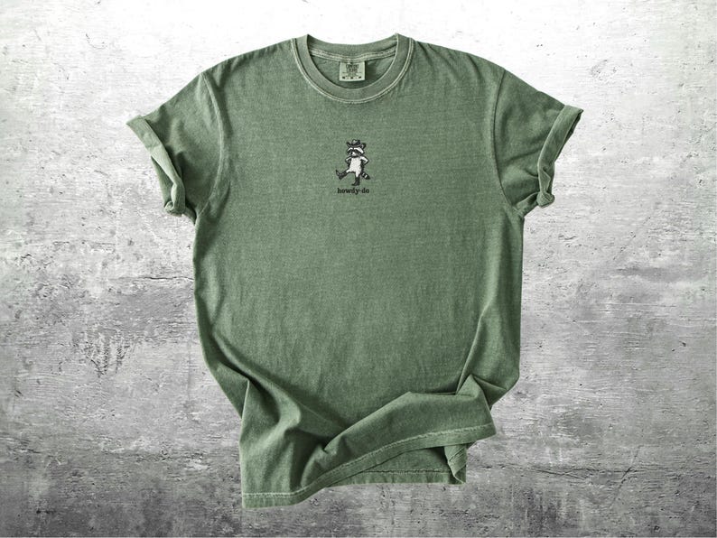May include: Olive green t-shirt with a small graphic of a raccoon wearing a hat and the text "howdy-do". The shirt has rolled-up sleeves and a crew neck. The fabric appears soft and slightly faded.