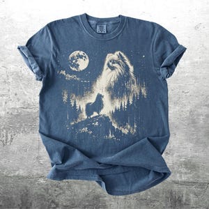 May include: Blue t-shirt featuring a graphic of a dog howling at the moon in a forest setting. The design includes a full moon, trees, and stars, creating a night scene. The shirt is made of soft material.
