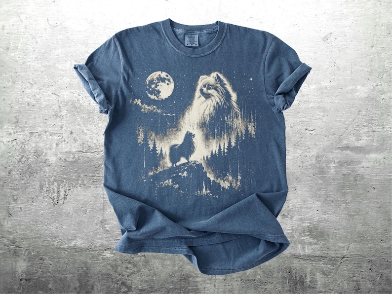 May include: Blue t-shirt featuring a graphic of a dog howling at the moon in a forest setting. The design includes a full moon, trees, and stars, creating a night scene. The shirt is made of soft material.