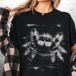 Op de afbeelding: Zwart T-shirt met een witte, verweerde spinnenafbeelding. Het spinnenontwerp heeft een getextureerde, geschetste uitstraling. Het shirt wordt gedragen met een zwart-beige geruit flanellen overhemd.