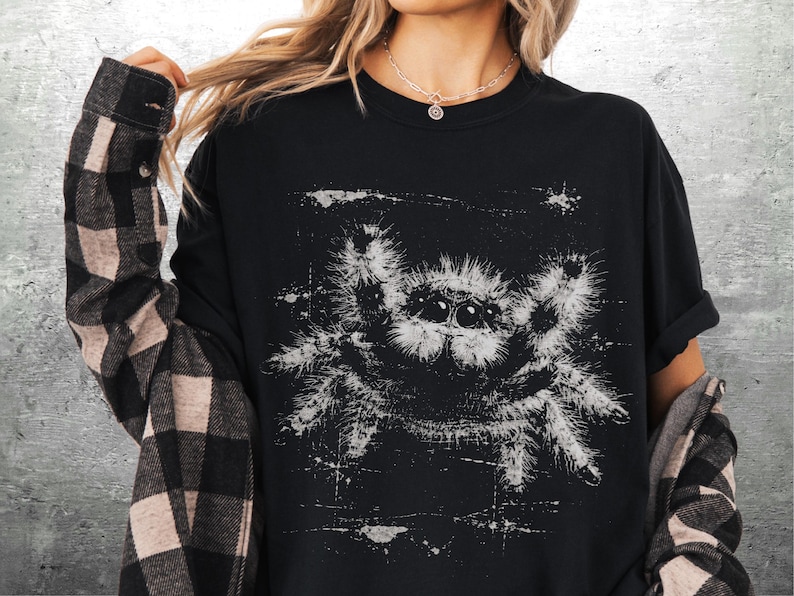 Op de afbeelding: Zwart T-shirt met een witte, verweerde spinnenafbeelding. Het spinnenontwerp heeft een getextureerde, geschetste uitstraling. Het shirt wordt gedragen met een zwart-beige geruit flanellen overhemd.