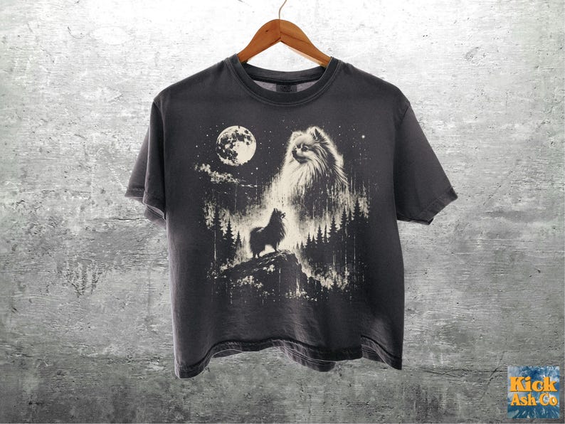 May include: A dark gray short-sleeve t-shirt featuring a graphic print of a fluffy dog standing on a rocky cliff in a forest at night under a full moon. The design includes a large ghostly dog face in the sky above the trees and the text Kick Ash-Co.