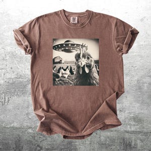 May include: Brown t-shirt with a sepia-toned graphic of a chicken looking at the viewer, with a UFO hovering above a barn and other chickens. The shirt has a crew neck and short sleeves.