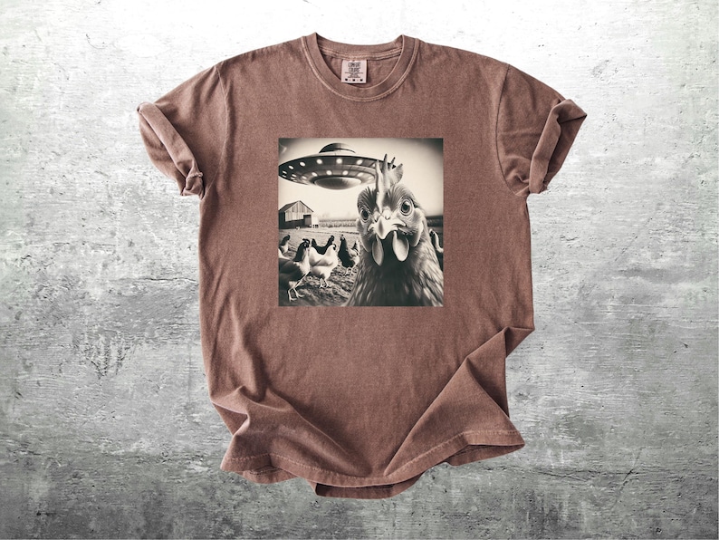 May include: Brown t-shirt with a sepia-toned graphic of a chicken looking at the viewer, with a UFO hovering above a barn and other chickens. The shirt has a crew neck and short sleeves.