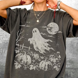 May include: A charcoal gray short-sleeve t-shirt featuring a distressed white graphic print of a ghost floating above a pumpkin patch with a full moon in the background. The design includes a bare tree and a wooden fence, with the text Kick Ash Co.