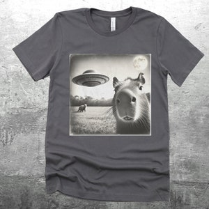 K&ouml;nnte beinhalten: Ein dunkelgraues T-Shirt mit einem Schwarzwei&szlig;bild eines Capybaras, das zu einem fliegenden Untertasse aufblickt. Das Bild ist im Vintage-Stil mit einem Mond im Hintergrund.