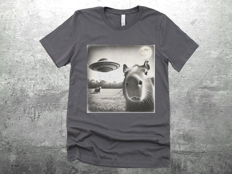 K&ouml;nnte beinhalten: Ein dunkelgraues T-Shirt mit einem Schwarzwei&szlig;bild eines Capybaras, das zu einem fliegenden Untertasse aufblickt. Das Bild ist im Vintage-Stil mit einem Mond im Hintergrund.