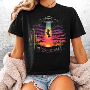 Puede incluir: Una camiseta negra con un estampado gr&aacute;fico de un perro salchicha siendo abducido por un platillo volante en un paisaje des&eacute;rtico al atardecer. La escena incluye cactus saguaro, un rayo de luz azul brillante y un cielo de tonos violetas y naranjas.
