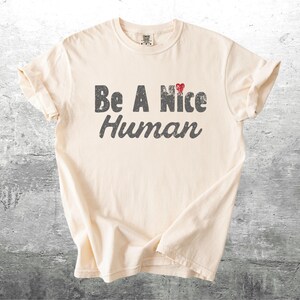K&ouml;nnte beinhalten: Cremefarbenes T-Shirt mit dem Aufdruck "Be A Nice Human" in verblichener grauer Schrift. Ein kleines rotes Herz akzentuiert das Wort "Nice". Das Shirt ist gefaltet und zeigt sein Design vor einem neutralen Hintergrund.