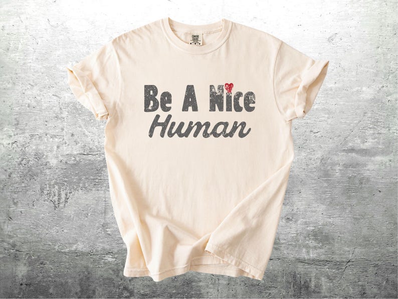 K&ouml;nnte beinhalten: Cremefarbenes T-Shirt mit dem Aufdruck "Be A Nice Human" in verblichener grauer Schrift. Ein kleines rotes Herz akzentuiert das Wort "Nice". Das Shirt ist gefaltet und zeigt sein Design vor einem neutralen Hintergrund.