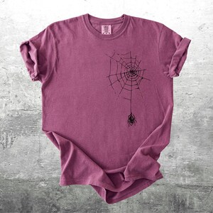 May include: A maroon t-shirt featuring a black spider web design with a spider hanging below. The t-shirt has rolled-up sleeves and a crew neck. The shirt is displayed on a gray concrete surface.