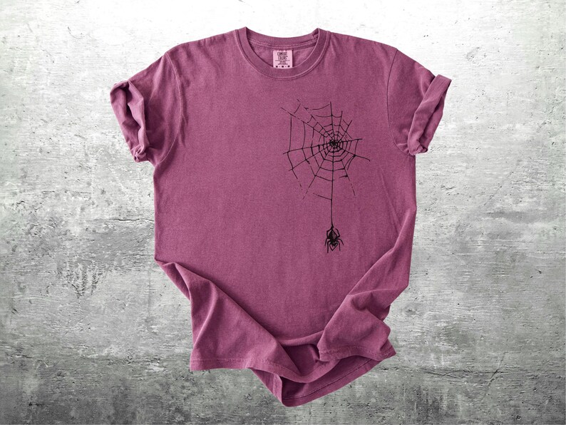 May include: A maroon t-shirt featuring a black spider web design with a spider hanging below. The t-shirt has rolled-up sleeves and a crew neck. The shirt is displayed on a gray concrete surface.