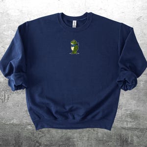 Puede incluir: Sudadera azul marino con un peque&ntilde;o bordado de una rana con una bufanda azul. La rana es verde con detalles amarillos. La sudadera tiene mangas largas y cuello redondo. El fondo es una superficie gris texturizada.