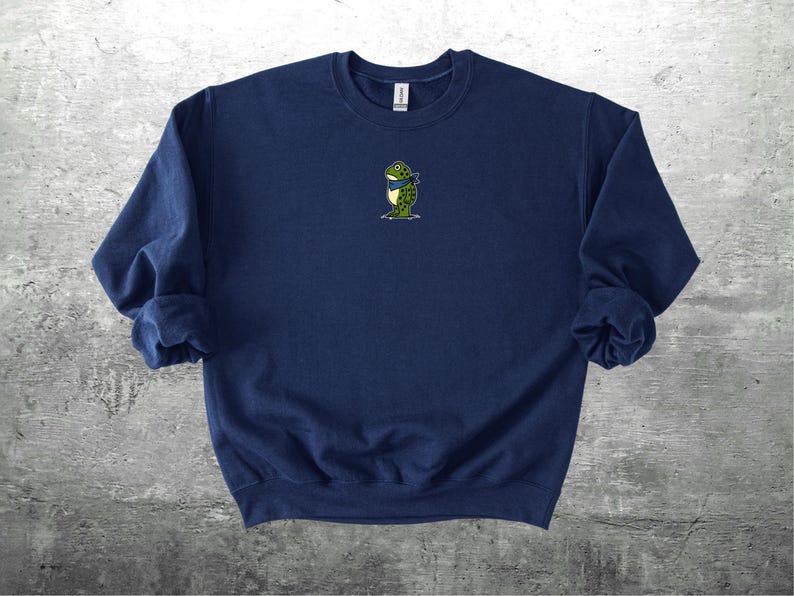 Puede incluir: Sudadera azul marino con un peque&ntilde;o bordado de una rana con una bufanda azul. La rana es verde con detalles amarillos. La sudadera tiene mangas largas y cuello redondo. El fondo es una superficie gris texturizada.