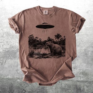 May include: Brown t-shirt featuring a black and white graphic of a capybara standing near a body of water, with a UFO above. The design includes trees and the text "Kick Ash Co."