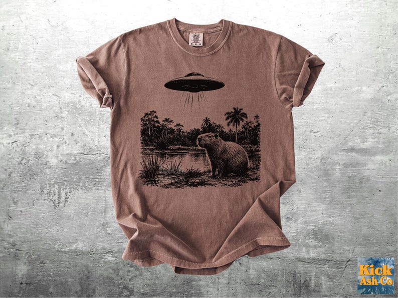 May include: Brown t-shirt featuring a black and white graphic of a capybara standing near a body of water, with a UFO above. The design includes trees and the text "Kick Ash Co."