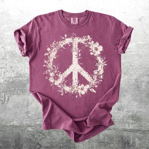 May include: A short-sleeved crew neck t-shirt in a muted berry-purple color. The center of the shirt features a large, off-white graphic of a peace sign surrounded by a detailed floral wreath pattern. The fabric has a slightly weathered texture.